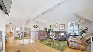 Un salón amplio con suelo de madera, alfombra verde, sofás de cuero, una mesa de centro de vidrio y un comedor con mesa y sillas de vidrio.