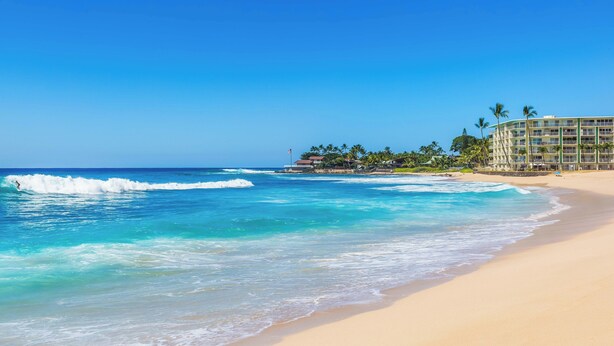 A beach with clear blue water, a sandy shore, and a resort building with multiple floors and palm trees. - Ocean Views Serene Vacation Escape Home Near Beach w/Beach Equipment & Games (Waianae)