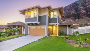 A two-story house with a garage, a well-maintained lawn, and a mountain backdrop. - Ocean Views Serene Vacation Escape Home Near Beach w/Beach Equipment & Games (Waianae)