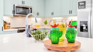 A kitchen with light blue cabinets, a stainless steel microwave, and a refrigerator with a black sign. There are two green cactus-shaped containers with yellow drinks and a metal basket with green plants. - Ocean Views Serene Vacation Escape Home Near Beach w/Beach Equipment & Games (Waianae)