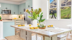 A modern kitchen with teal cabinets, a wooden dining table set with yellow napkins, and a view of a mountainous landscape through large windows. - Ocean Views Serene Vacation Escape Home Near Beach w/Beach Equipment & Games (Waianae)
