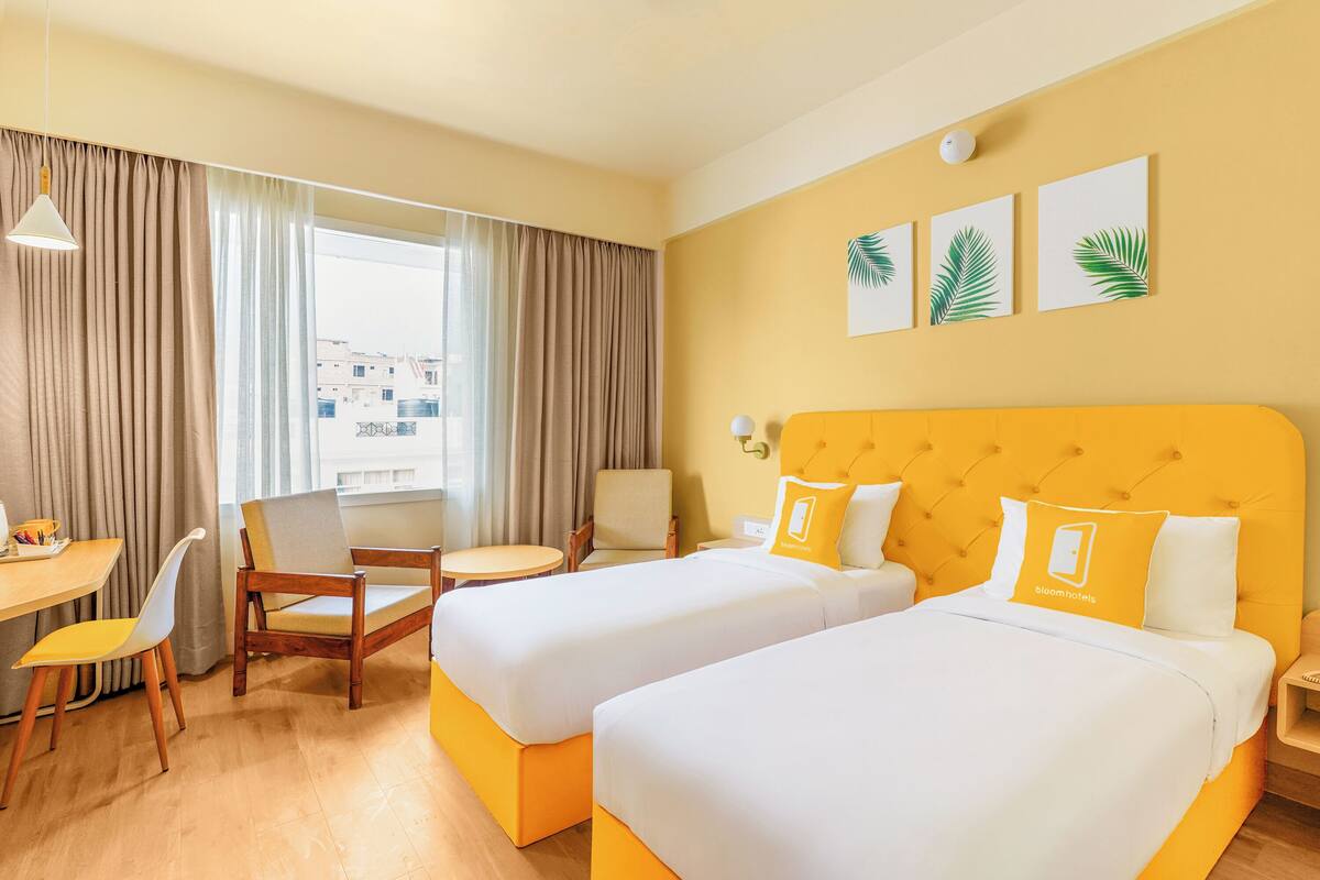 A hotel room with two beds, a desk, chairs, and a view of buildings through the window.