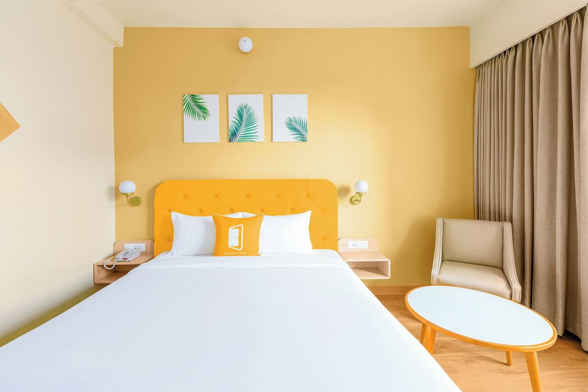 A hotel room with a large bed, a bedside table with a lamp, a chair, and a small table. There are three wall-mounted artworks above the bed.