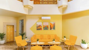 Un vestíbulo amarillo y luminoso con un sofá, dos sillones, otomanas, un mostrador de recepción y plantas en macetas.