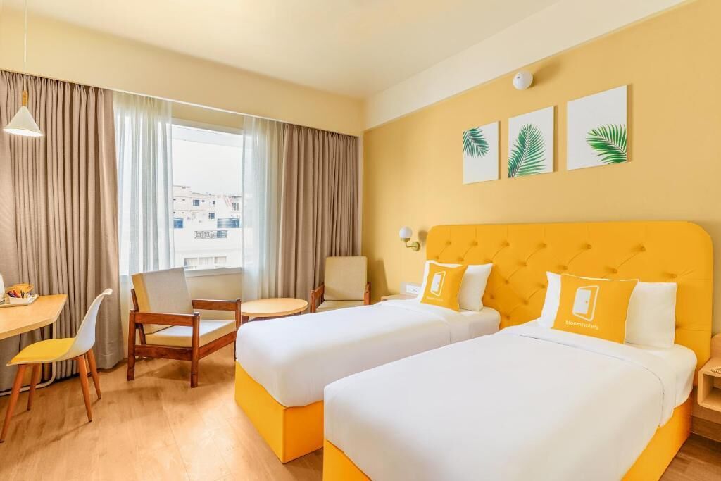 A hotel room with two beds, a dining table, a chair, a small table, and a window with curtains.