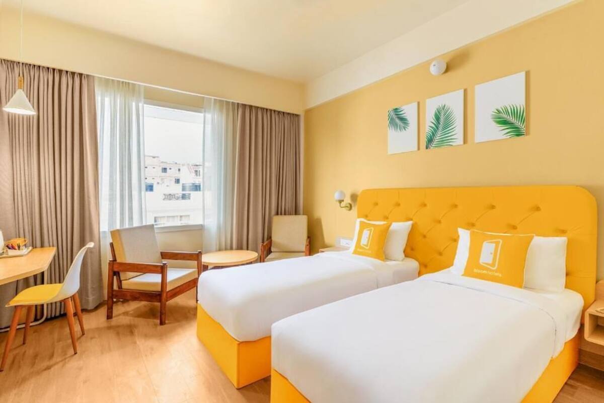 A hotel room with two beds, a dining table, a chair, a small table, and a window with curtains.