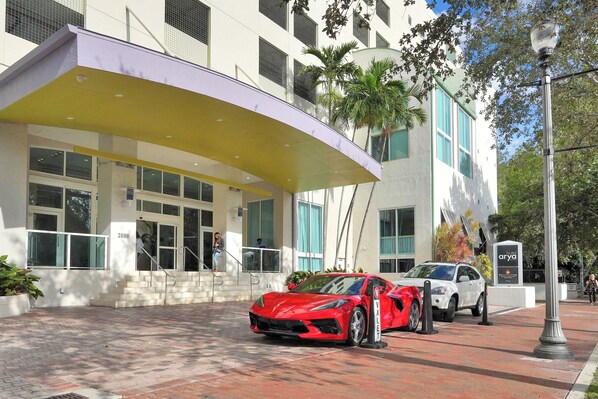 A hotel entrance with a red sports car parked in front, a white car nearby, and a covered walkway leading to the entrance. - Stunning 12th Floor Studio in the Grove (Miami)