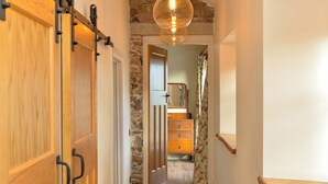 A narrow hallway with wooden doors and stone walls, leading to a staircase with a wooden handrail and a hanging light fixture.