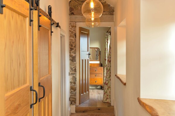 Un couloir étroit, avec des portes en bois et des murs en pierre, qui mène à un escalier doté d’une rampe en bois et d’un luminaire suspendu.