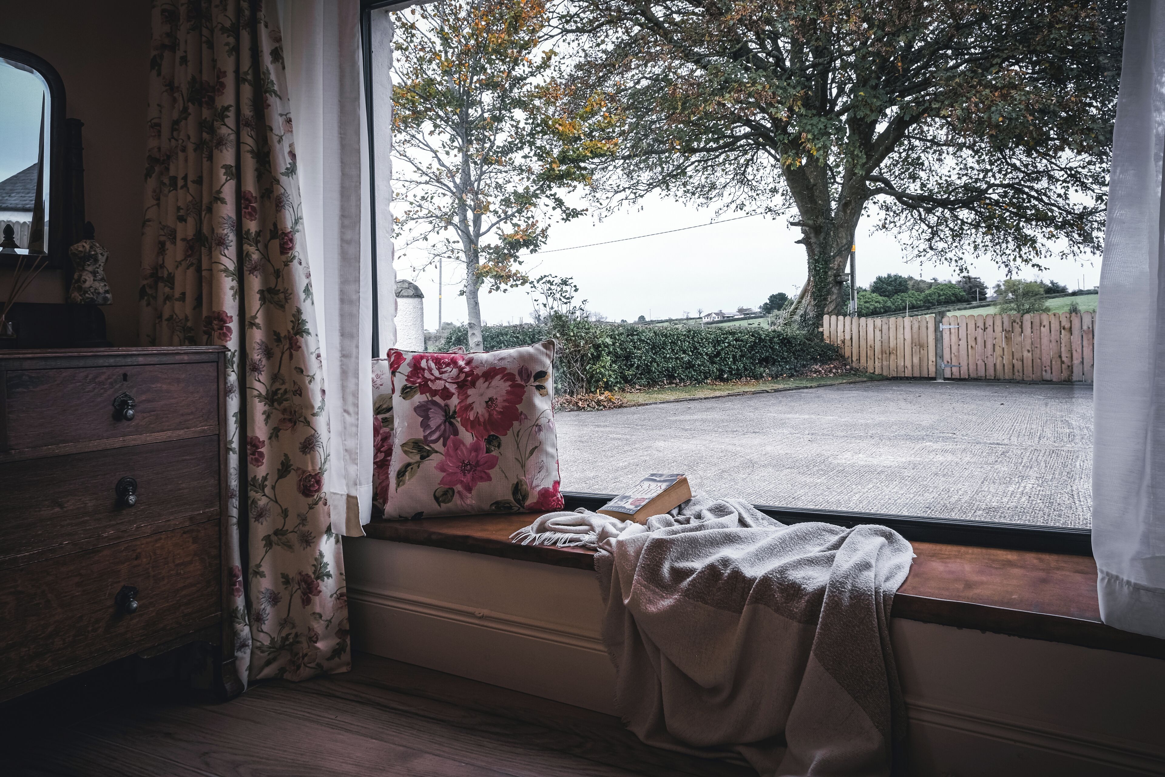 Une pièce avec un siège de fenêtre, une commode en bois, un coussin à motifs floraux et une couverture.