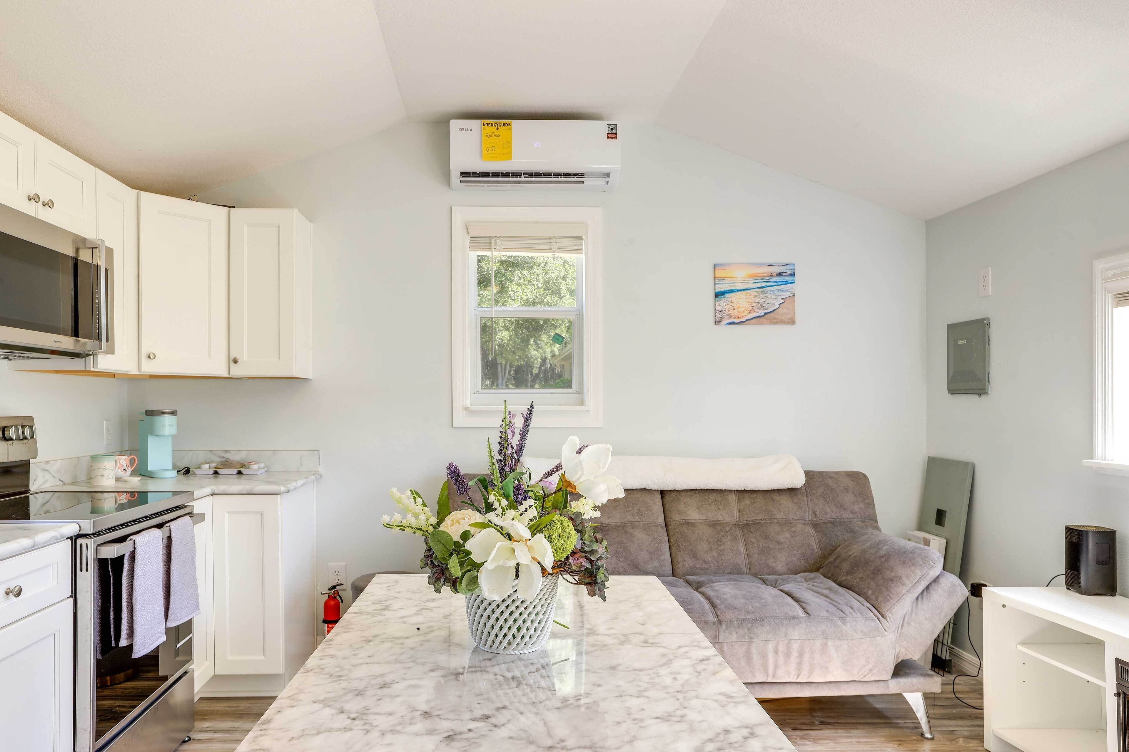 A modern kitchen with white cabinets, a microwave, and a coffee maker. There is a marble countertop with a vase of flowers. A gray sofa and a white table are visible in the living area.