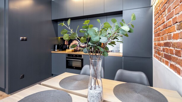 Una cocina moderna con armarios oscuros, una pared decorada con ladrillos y una zona de comedor con una mesa de madera y posavasos grises.