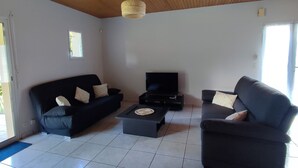 Una sala de estar con dos sofás negros, una mesa de centro y un televisor de pantalla plana sobre un mueble bajo. El techo es de madera y las paredes son blancas.