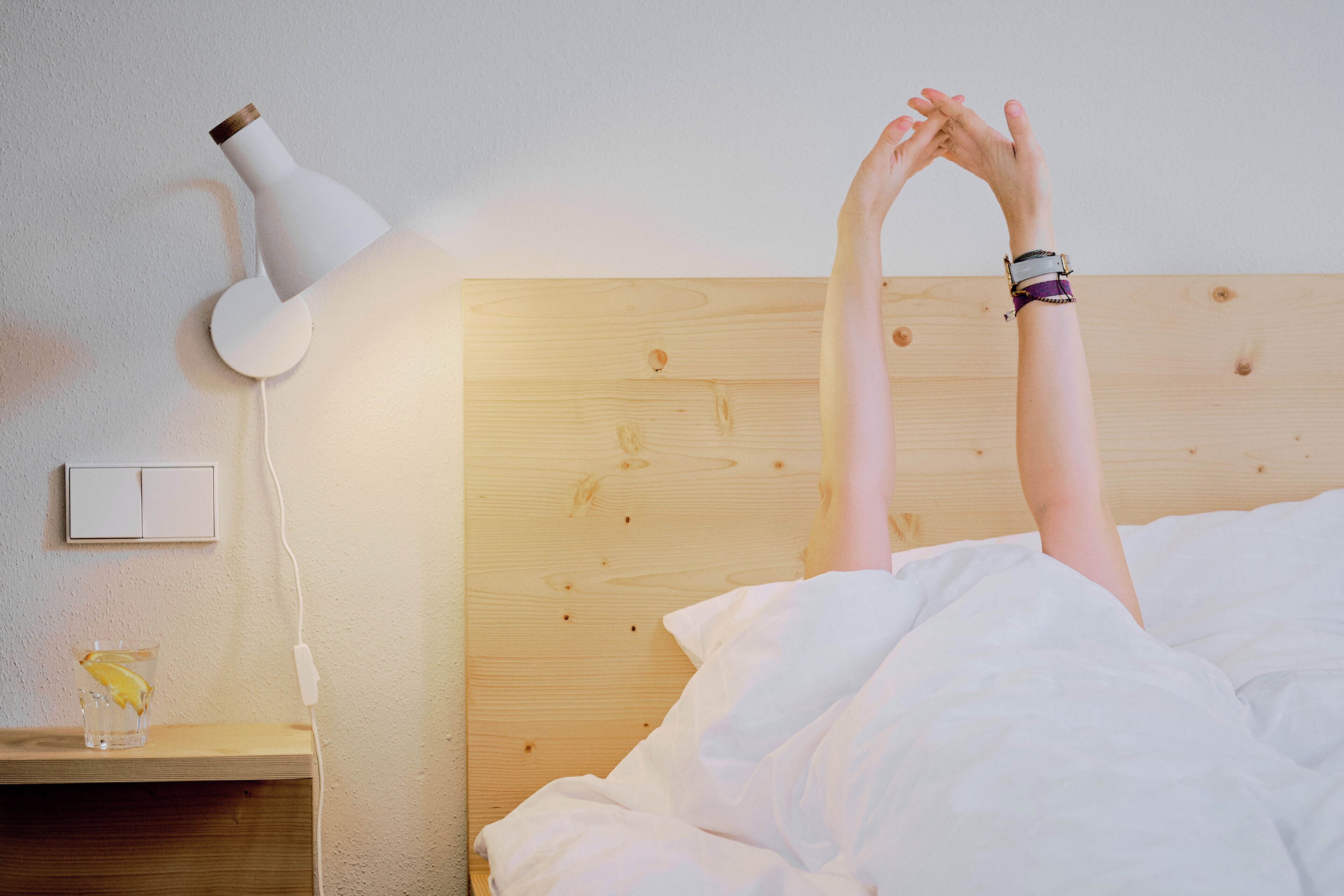 Una persona acostada en una cama con ropa blanca, un cabecero de madera, una lámpara de noche blanca y un enchufe en la pared.