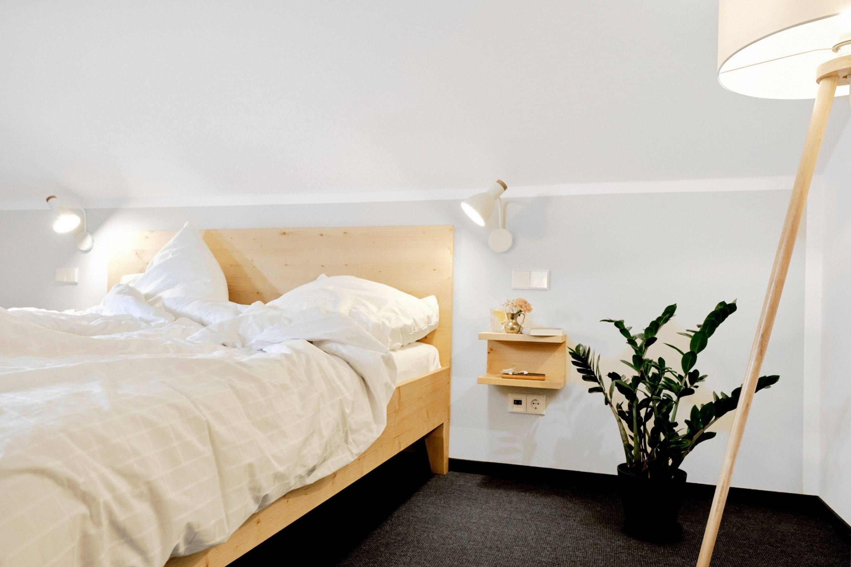 Ein ordentlich bezogenes Bett mit weißen Leinen, ein Holz-Kopfbrett, ein Nachttisch mit Lampe und eine Topfpflanze.