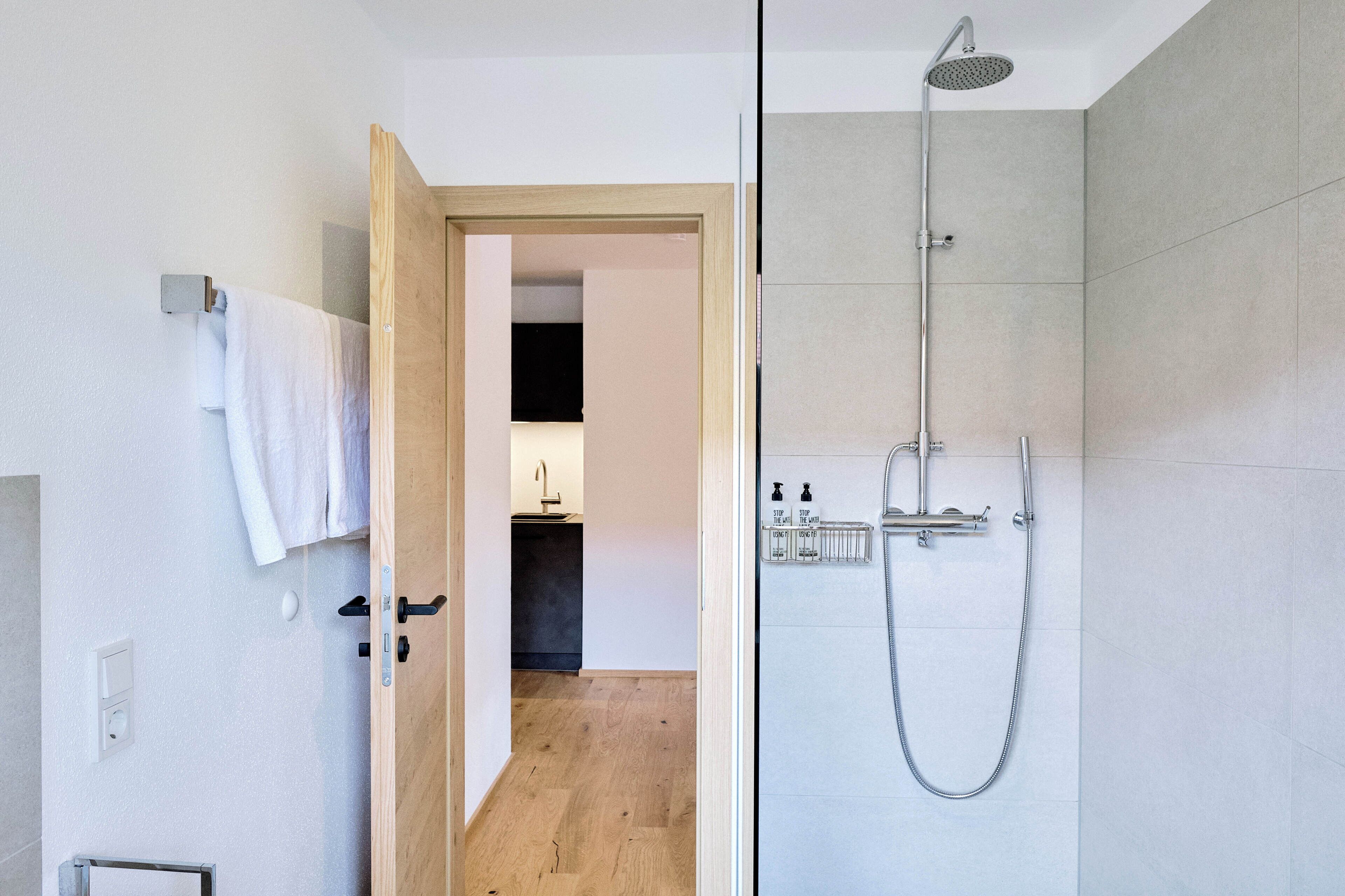 Un baño moderno con ducha de vidrio, una puerta de madera que da a un pasillo y un perchero visible.