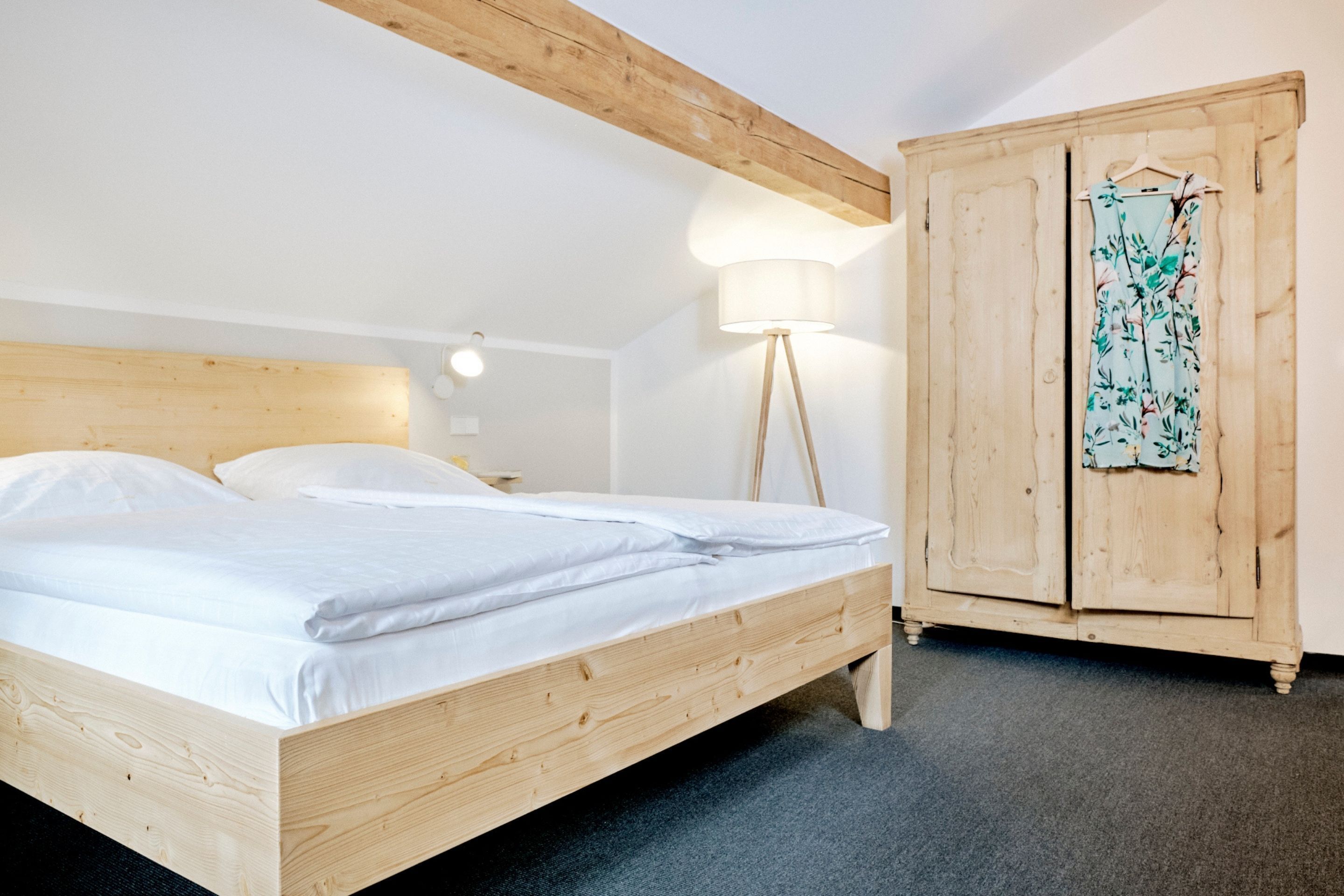 Een houten bed met witte lakens, een houten kledingkast en een vloerlamp in een kamer met witte wanden en een houten plafondbalk.