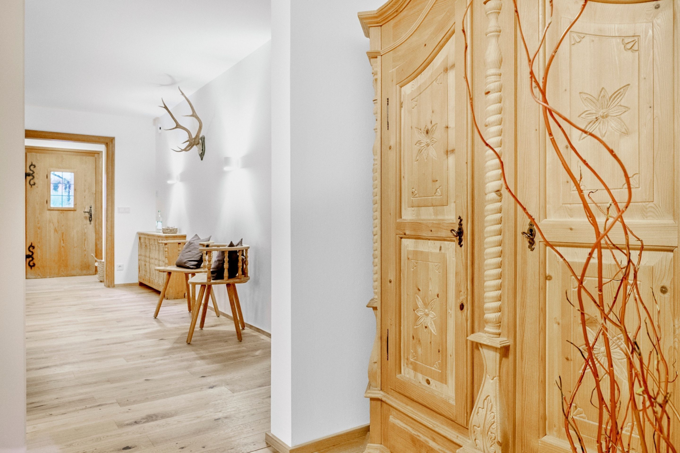 Un couloir doté d’un plancher en bois, d’une porte en bois, d’un meuble orné de motifs sculptés et d’une paire de bois de cerf fixée au mur.