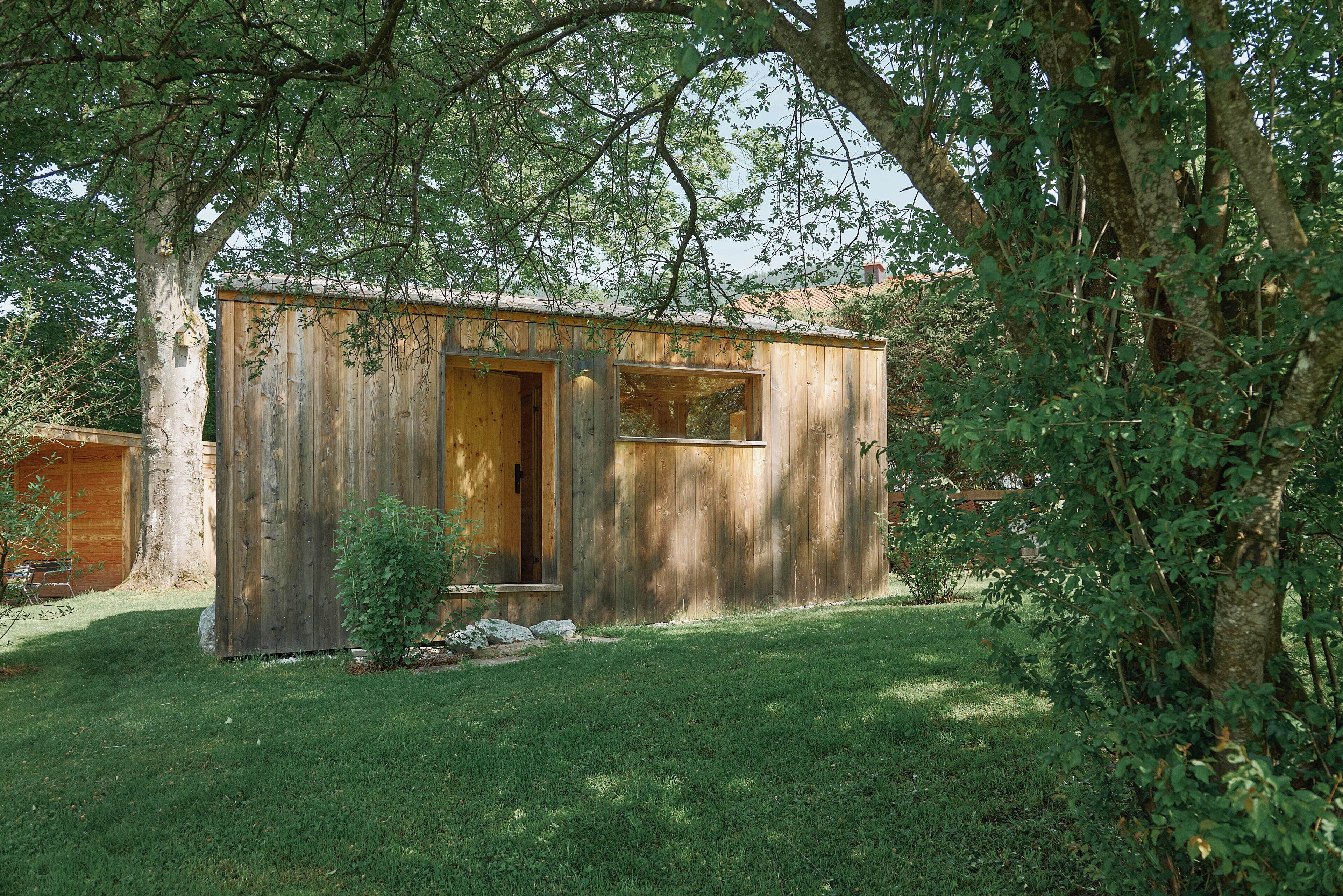 A small wooden cabin with a door and two windows, surrounded by trees and grass.