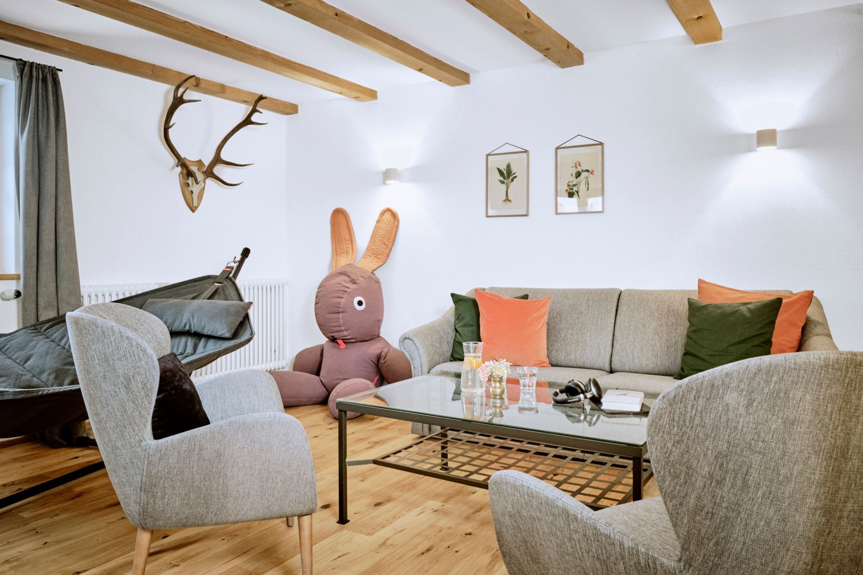 A modern living room with a sofa, chairs, a glass coffee table, a teddy bear, and wall-mounted deer antlers.