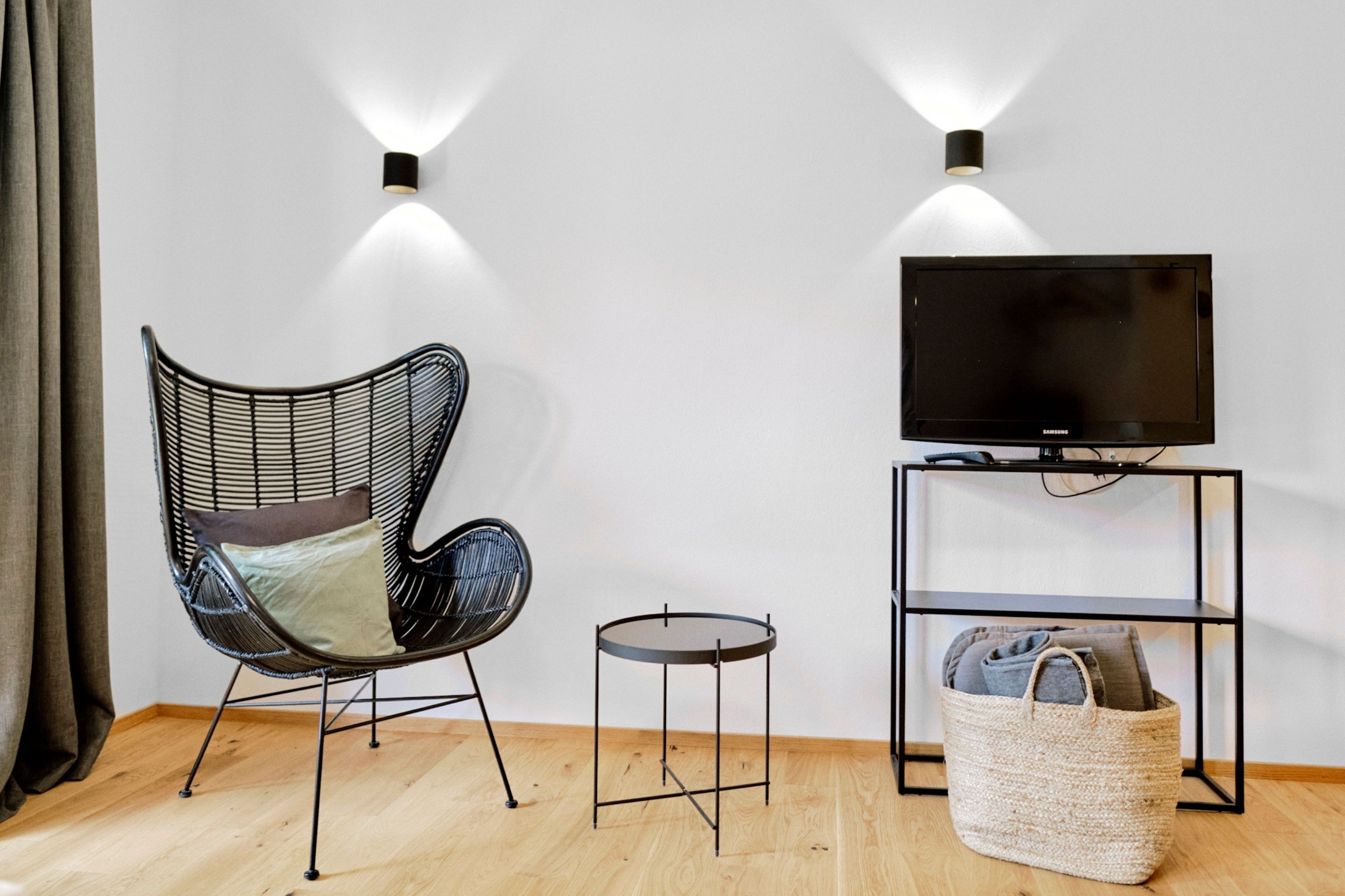 Une pièce moderne avec une chaise noire en osier, une petite table ronde, un téléviseur posé sur un meuble noir et un panier en osier sur le sol.