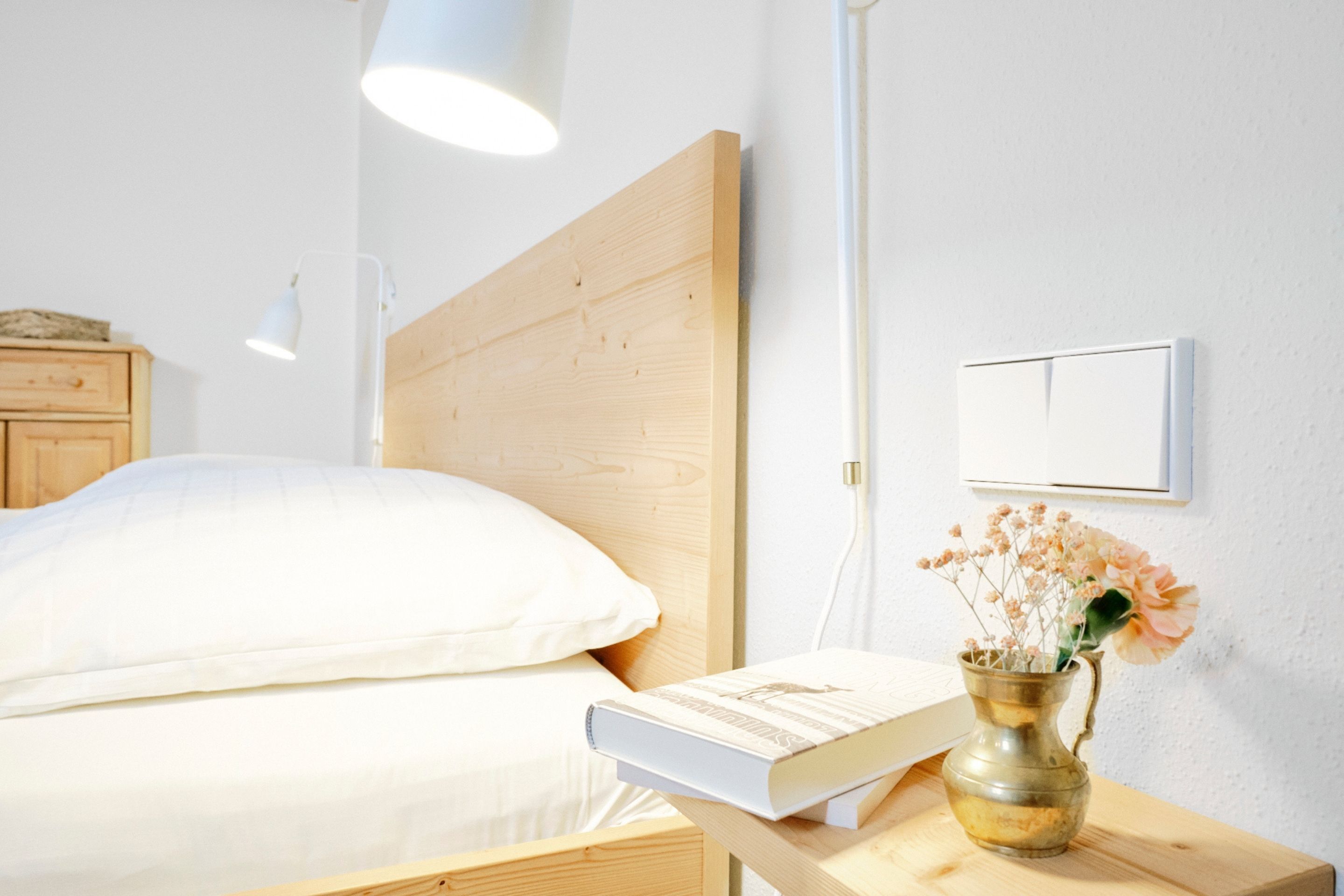 Un letto rifatto con un cuscino bianco e un libro accanto. Su un comodino in legno, una brocca di ottone con dei fiori.
