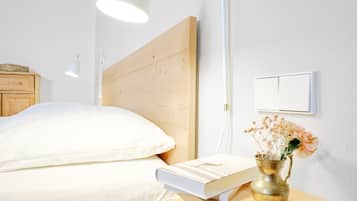 Un letto rifatto con un cuscino bianco e un libro accanto. Su un comodino in legno, una brocca di ottone con dei fiori.