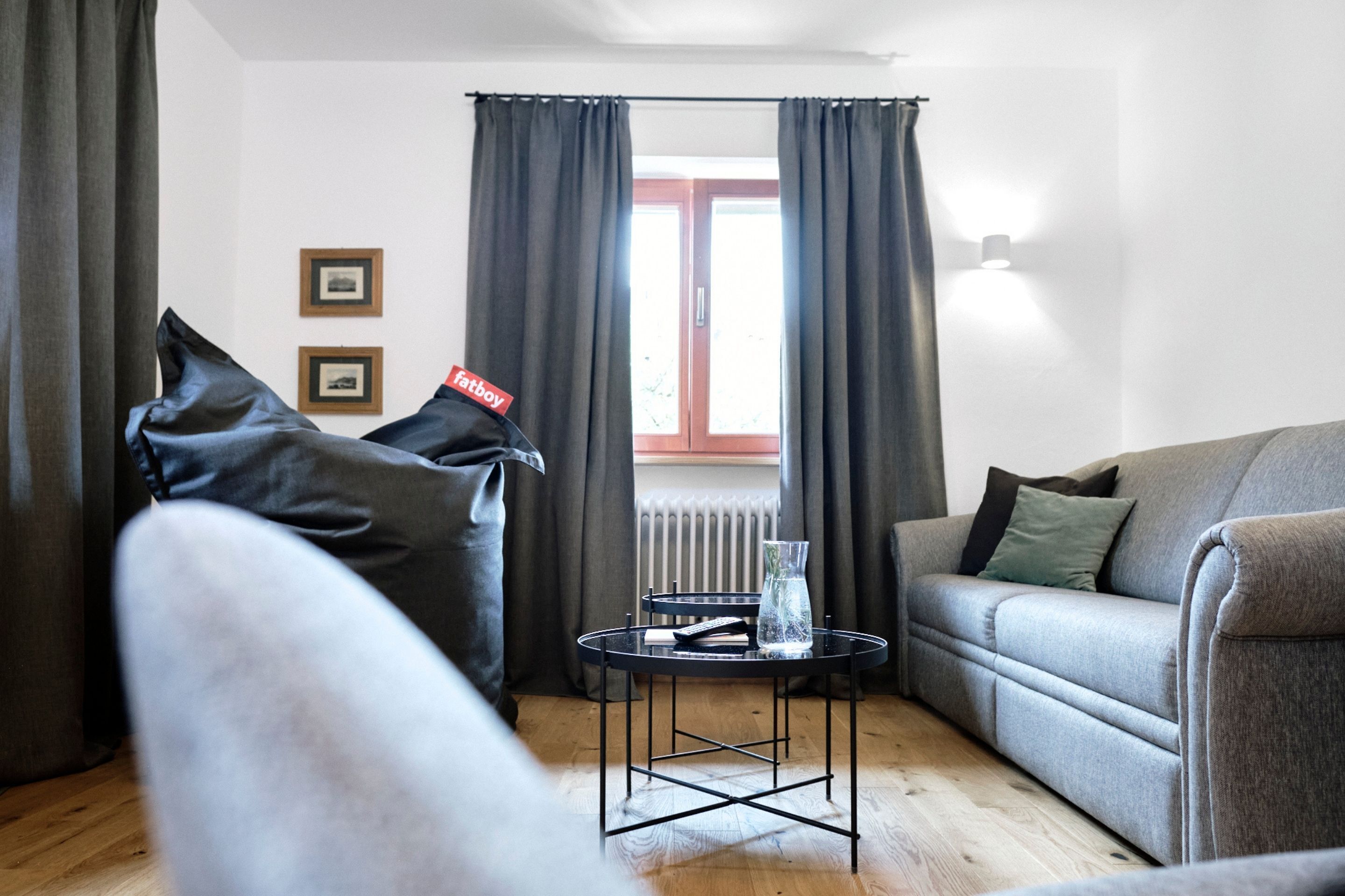 A modern living room with a grey sofa, a small round table with a vase, and a view of a window with curtains.