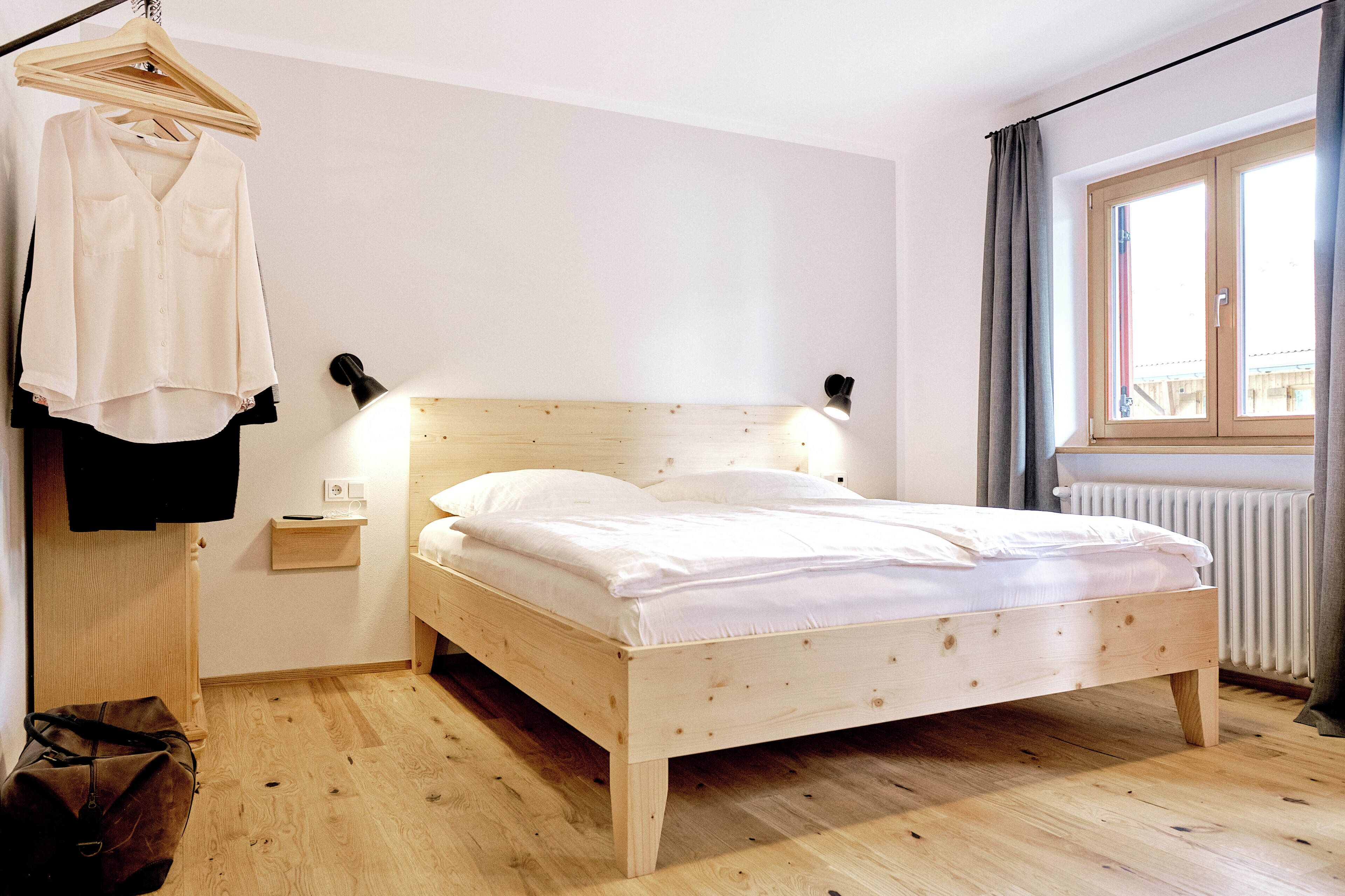 A wooden bed in a room with a hanging clothes rack, a bedside table, and a window with curtains.