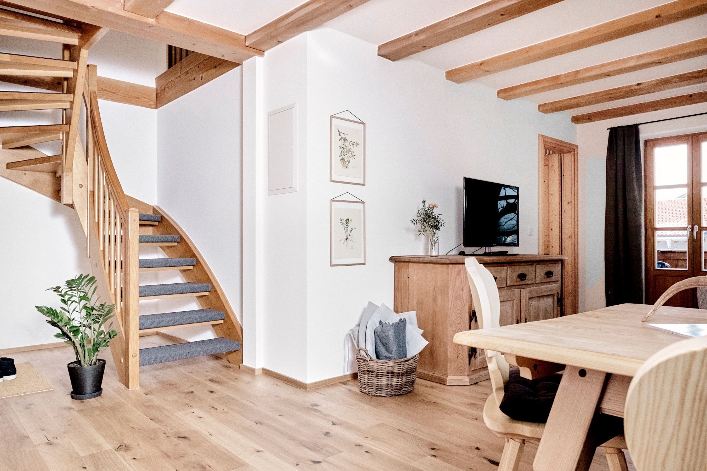 A wooden staircase, a dining table, and a TV in a room with white walls and wooden beams.