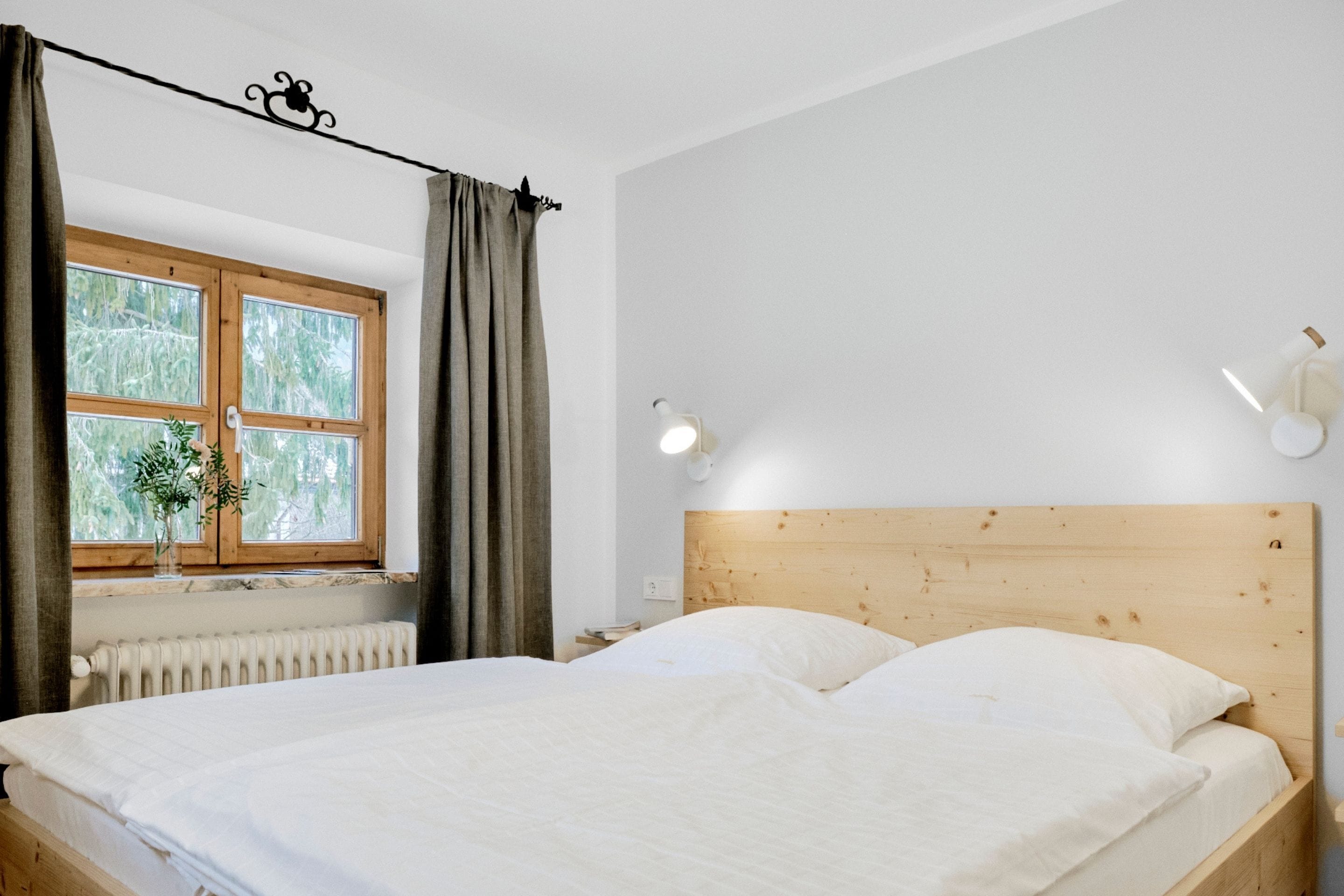 Un dormitorio con cabecera de madera, una cama grande con ropa de cama blanca, una ventana con cortinas, una mesita de noche con lámpara y un radiador.