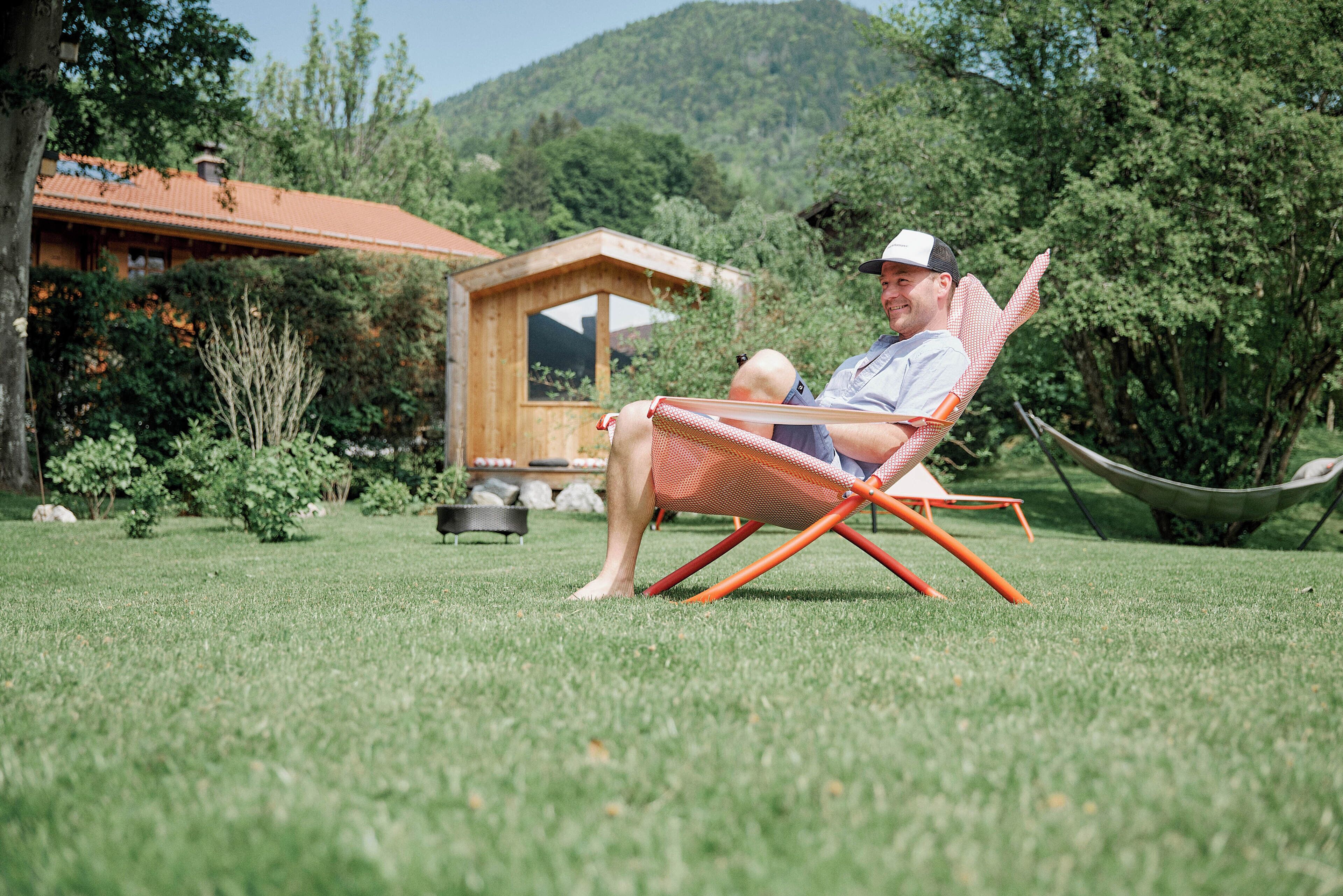Un hombre descansando en una silla naranja sobre un césped, con una cabaña de madera y árboles al fondo.