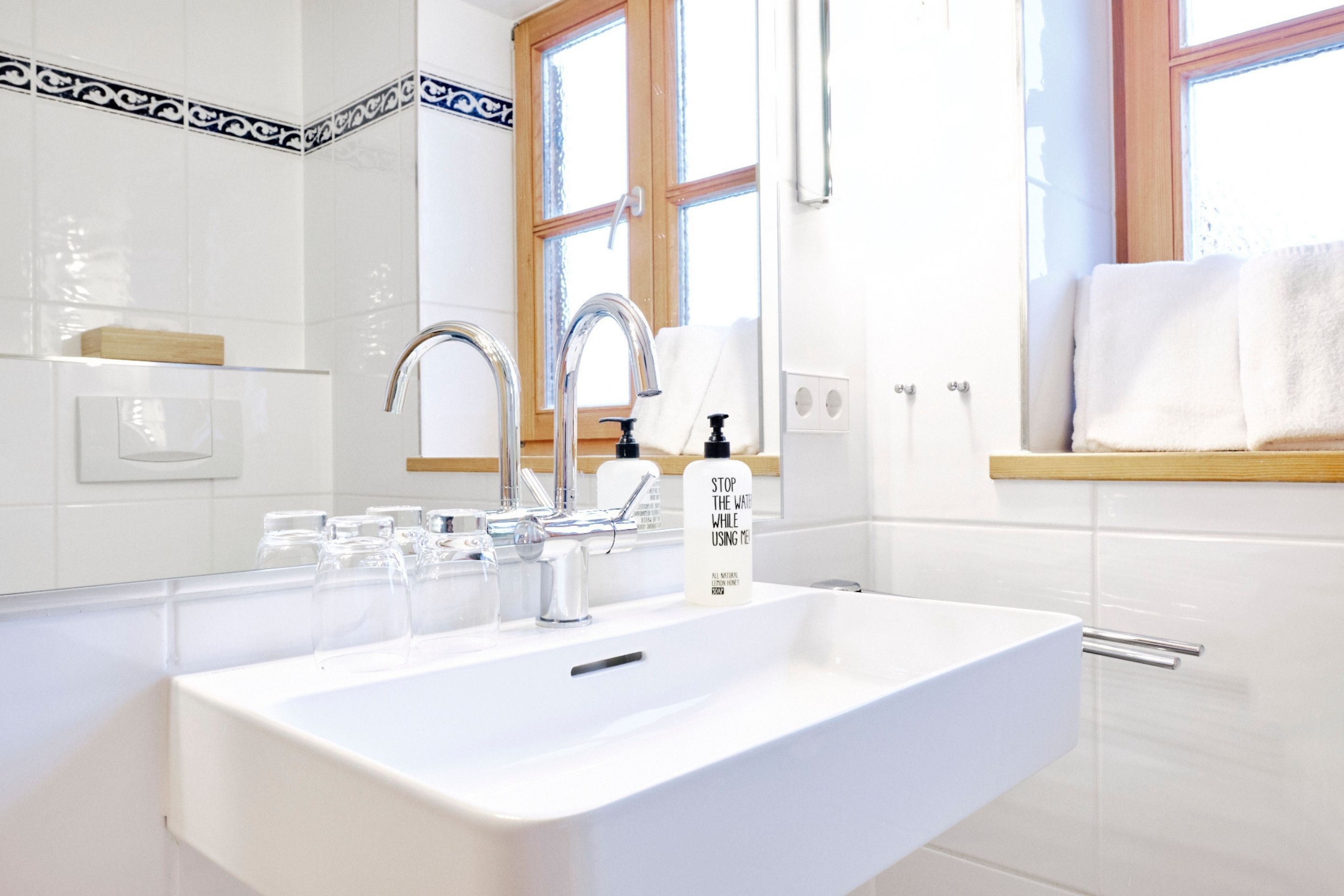 A modern bathroom with a white sink, two faucets, and a window above the sink.
