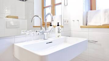 A modern bathroom with a white sink, two faucets, and a window above the sink.