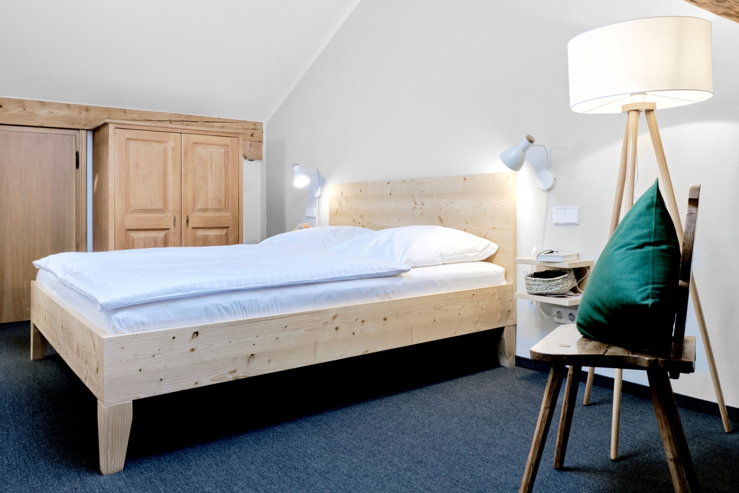 A single bed with a wooden frame and white bedding, a bedside lamp, a green pillow, a small table with a lamp, and a wooden wardrobe.