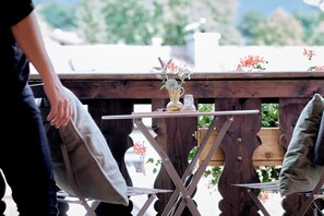 A wooden deck with a small table and chairs, a vase with flowers, and a person holding a pillow. - Der Lederer Hof (Tegernsee)