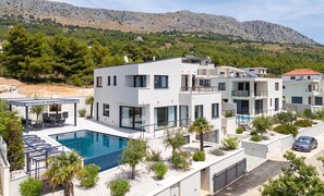 A modern multi-story residential building with a swimming pool, outdoor seating, and a view of a mountainous landscape. - Villa Apolina 4-en-suite bedrooms, heated 36sqm pool, sea views, beach 1km (Podstrana)