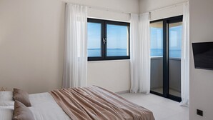 A bedroom with a bed, a large window with curtains, a sliding glass door to a balcony, and a flat-screen TV mounted on the wall.
