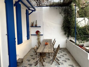 A wooden table with chairs in a white-walled outdoor area with blue accents, a tiled floor, and potted plants on shelves. - A HIDDEN GEM: A UNIQUE VACATION HAVEN IN THE HEART OF MALMOUSQUE (Marseille)