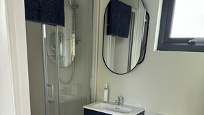 Un baño pequeño con cabina de ducha de vidrio, inodoro blanco, mueble oscuro de dos puertas y un espejo redondo en la pared.
