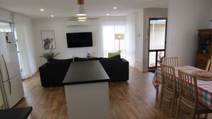 Una cocina moderna con una isla central, una zona de comedor con mesa y sillas, un sofá y un televisor de pantalla plana montado en la pared.