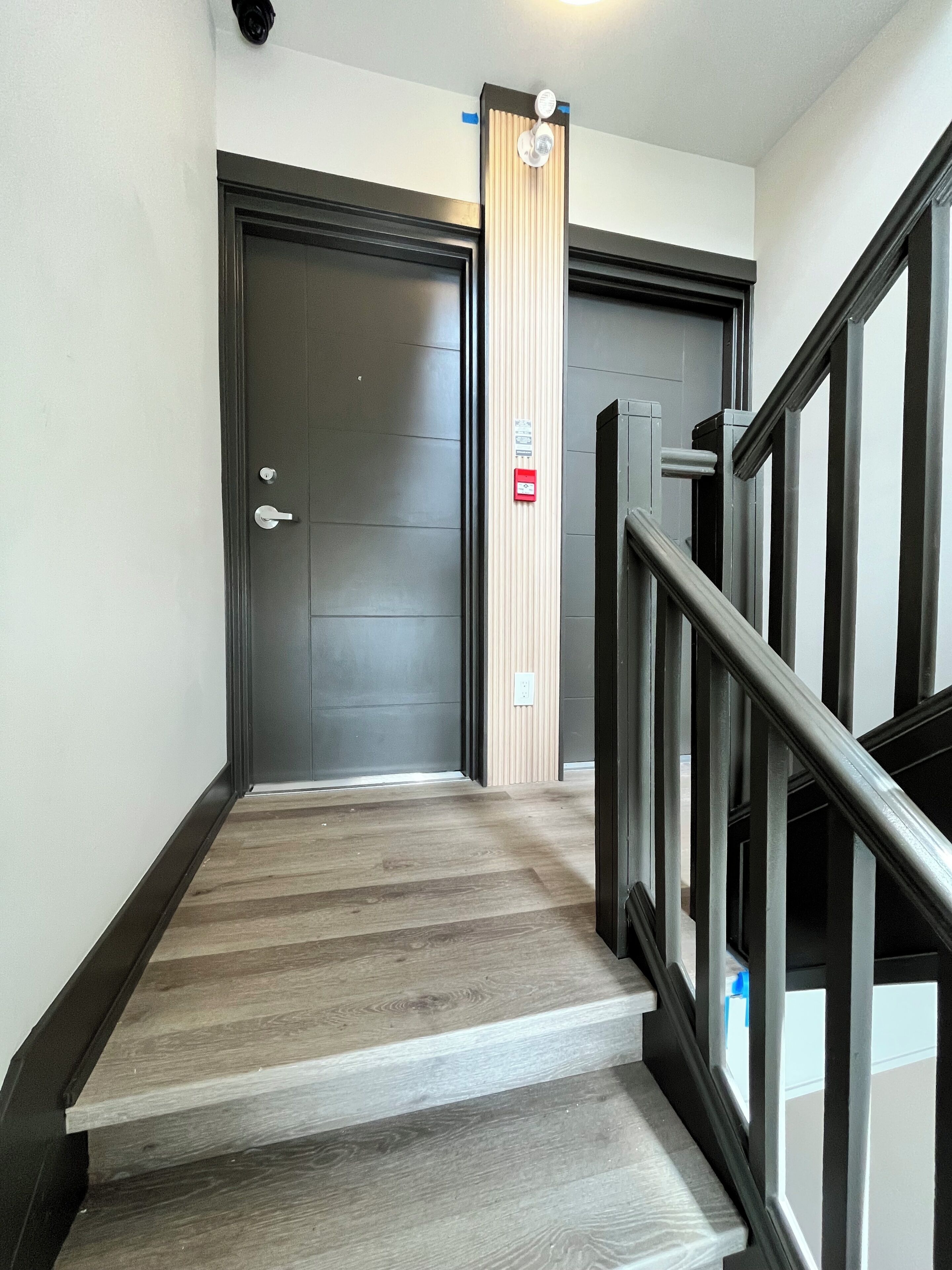 Un escalier avec des marches recouvertes de bois, une porte noire et un bouton d’alarme rouge.