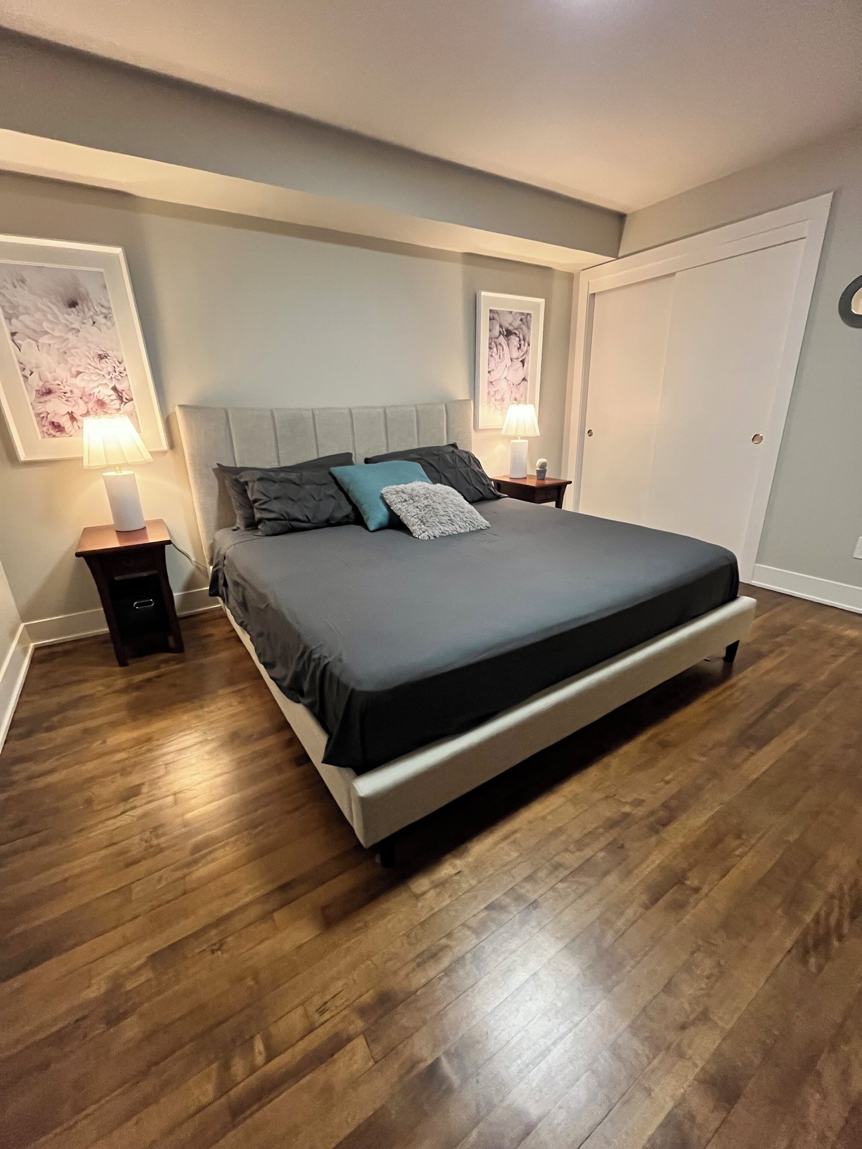 A bedroom with a large bed, two bedside tables with lamps, a wardrobe, and two framed floral artworks on the walls.