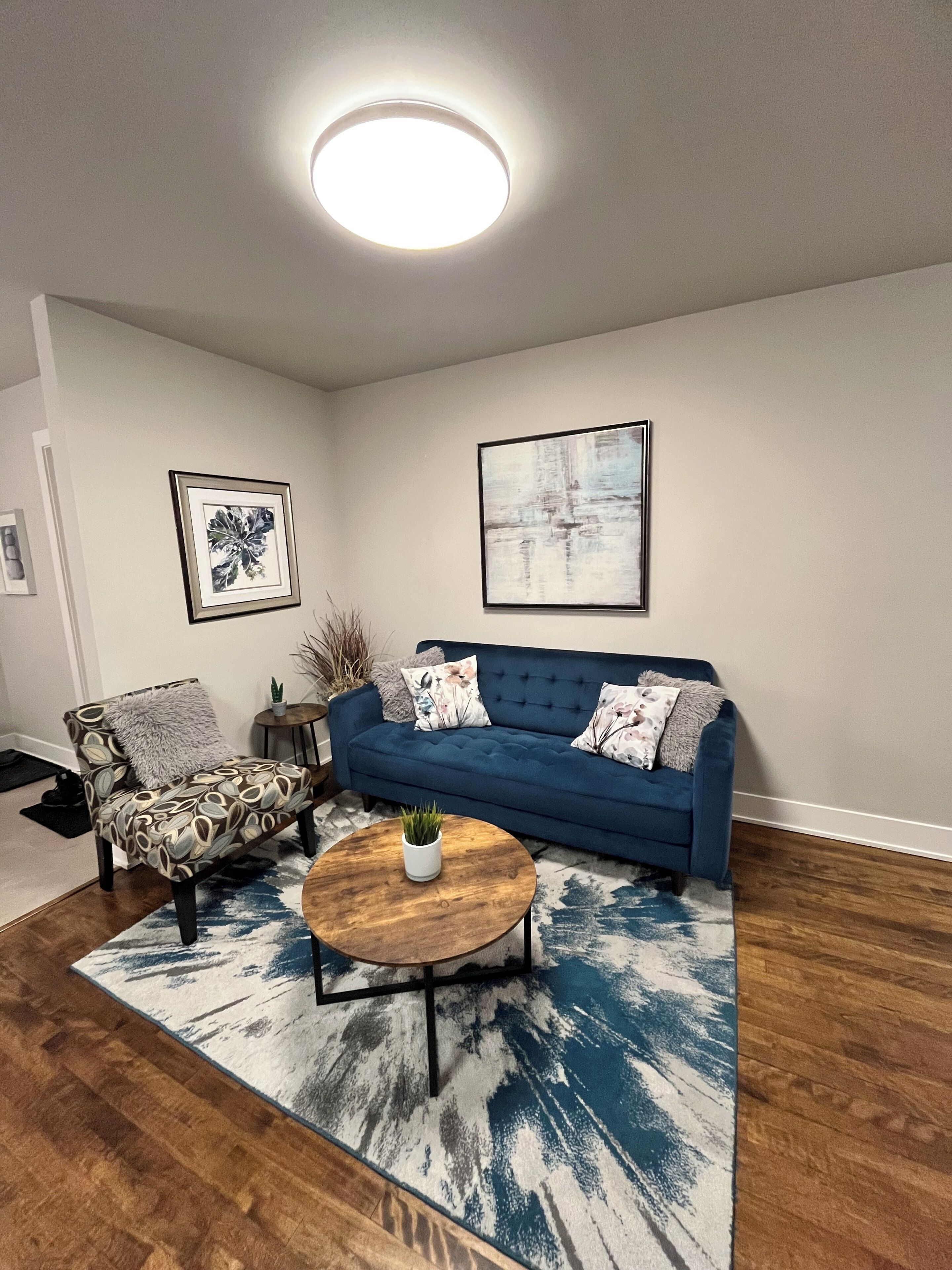 A living room with a blue sofa, a round coffee table, a patterned armchair, and two framed artworks on the wall.