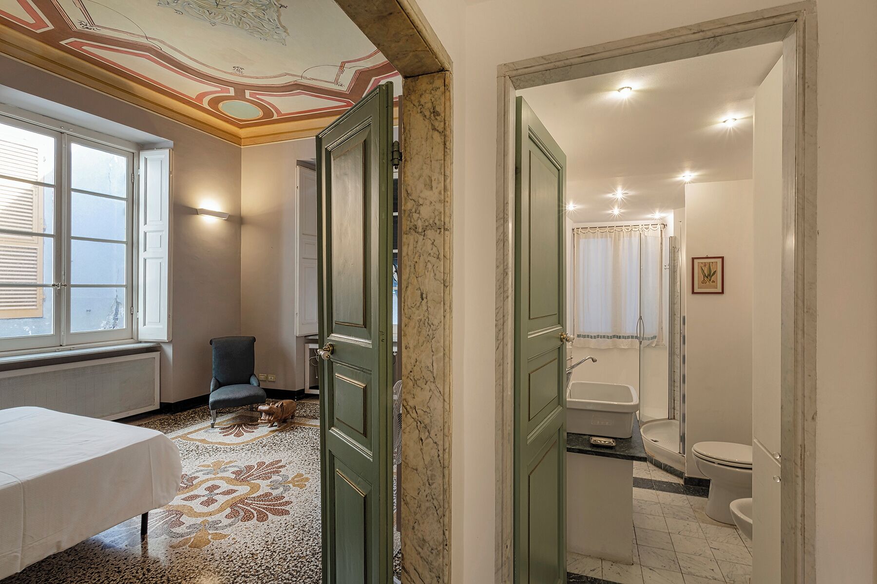A hotel room with a large window, a green door, a marble floor, a white bed, and a bathroom with a sink and toilet.