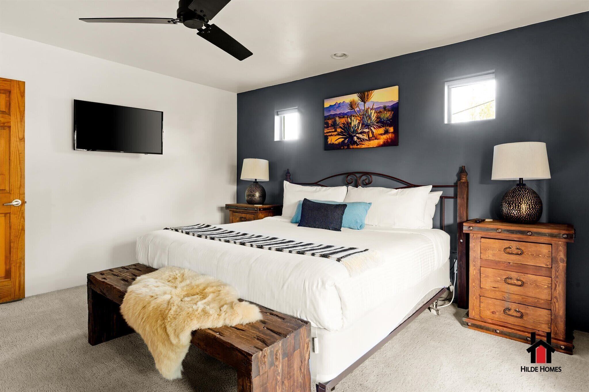 A bedroom with a bed, a ceiling fan, a TV, a painting, and a wooden nightstand with drawers.
