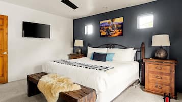 A bedroom with a bed, a ceiling fan, a TV, a painting, and a wooden nightstand with drawers.