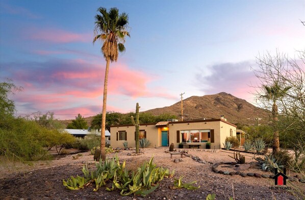 A desert-style house with a single-story structure, surrounded by cacti and desert plants, set against a backdrop of a mountain and a colorful sky.