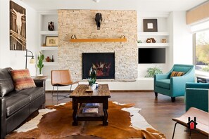 A modern living room with a stone fireplace, leather sofa, teal armchairs, and a wooden coffee table.