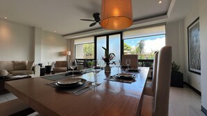 A modern dining area with a wooden table set for four, a ceiling fan, and large windows offering a view of greenery. - Serene Beachside 2 bedroom and 2.5 bathroom condo. (Playa Hermosa)